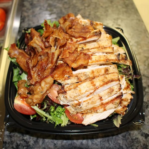 Grilled chicken and crispy bacon strips on a bed of fresh greens and tomato slices in a black container.